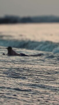 View of person enjoying water recreation at beach during sunset, lying prone on a surfboard or bodyboard, gentle ocean waves moving towards shore, soft light creating peaceful atmosphere. Vertical sho