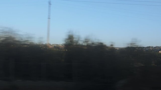 Dynamic view from moving train, showcasing fast-passing countryside scenery. Motion blur affects foreground trees and houses. Clear blue sky dominates upper frame. Distant village sits on a low hill, 