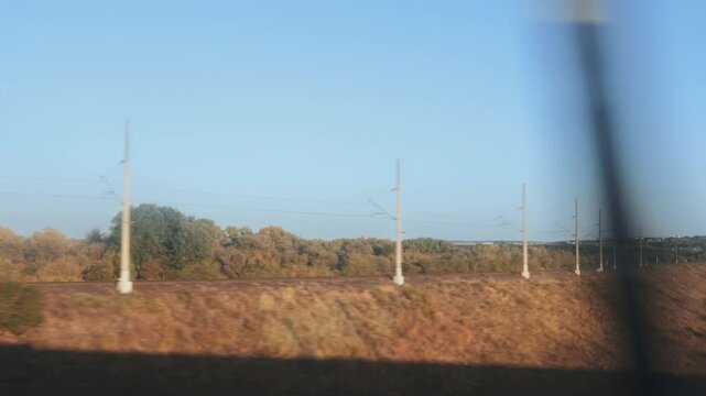 Scenic view from moving train, revealing extensive landscape of railway track, electric poles, and autumnal foliage. Dry golden grass covers side of embankment, leading to distant small town.