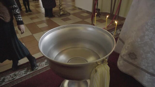 View of large metal baptismal font filled with water, positioned on red carpet in church interior with tiled floor. Lit candles burn near font, suggesting upcoming religious ceremony, people stand nea