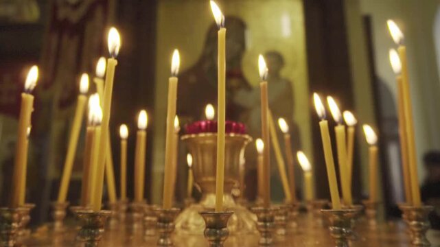 Many tall, slender, yellow wax candles flicker with vibrant flames, casting soft, warm glow on golden candlesticks and ornate altar within solemn church interior. Blurred background features ancient r