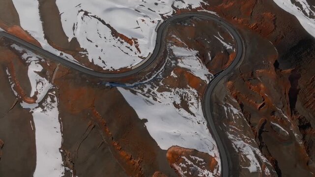 Drone footage looking straight down on a curving asphalt road crossing snowy mountain slopes at Chyyyrchyk Pass, Kyrgyzstan, with exposed brown rock and ridges