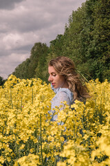 Fototapeta premium A young woman with wavy hair, wearing a light blue floral dress, stands in a vast field of bright yellow blooming rapeseed (canola) flowers. She poses with her hands behind her head, 