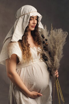 Serene Pregnant Woman in Medieval White Gown Holding Dried Flowers