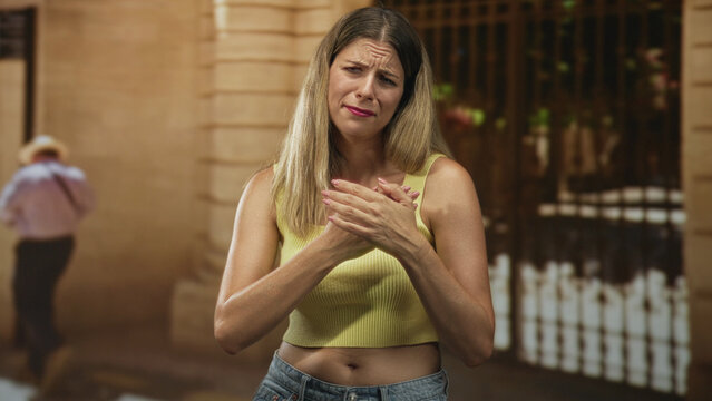 Woman clutching her chest with both hands on a street in front of a building facade wearing a yellow crop top and jeans; emotional distress vulnerability.