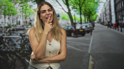 Fototapeta premium Woman with index finger to chin and glasses, smiling with arms crossed at waist on street near parked cars and bicycles; thoughtful curiosity.