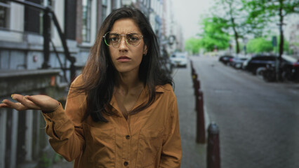 Fototapeta premium Woman shrugging with palms up on a city street wearing brown shirt and glasses, looking puzzled at an empty sidewalk; confusion questioning.