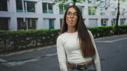 Fototapeta premium Woman wearing glasses smiles and turns head on a city street, long hair draped over shoulder, casual jeans and belt visible; confidence curiosity.