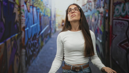 Naklejka premium Woman wearing white top and jeans raises hands palms up in a graffiti lined street, bare hands visible, standing between painted walls; frustration release resilience.