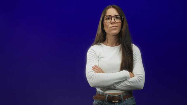 Woman with glasses, arms crossed, long hair, wearing white top, jeans and brown belt against a deep purple studio backdrop; quiet determination.