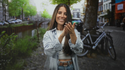 Naklejka premium Young hispanic woman clapping hands and smiling, bare midriff visible, standing on a street beside a canal with parked bicycle and trees; joy celebration.