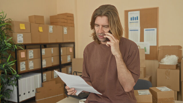 Man holding smartphone to ear reads a parcel invoice amid stacked cardboard boxes in a building; concern small business.