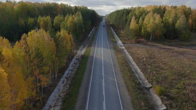 Drone footage of a long straight asphalt highway cutting through colorful autumn forest, with a single car driving in the distance, Ural region, Russia