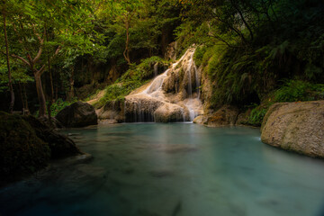 A turquoise waterfall cascades down in layers in the middle of the forest. © Pongsorn