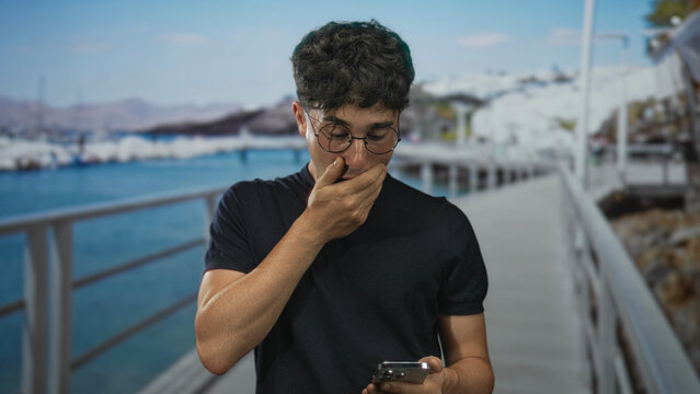 Young man on seaside promenade holding smartphone with hand covering mouth while looking down at the screen; surprise uncertainty.