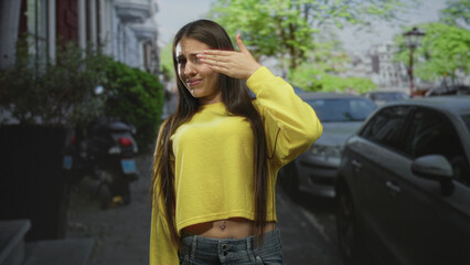 Fototapeta premium Young woman covering eyes with hand on street with parked cars, cropped yellow sweater revealing navel piercing and denim jeans; shyness privacy.