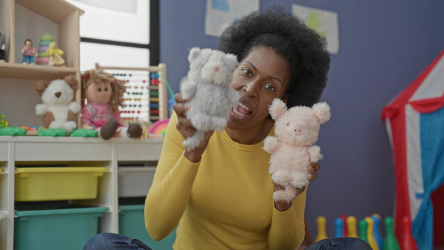Woman holding two plush hand puppets while speaking in a colorful preschool classroom; playful learning.