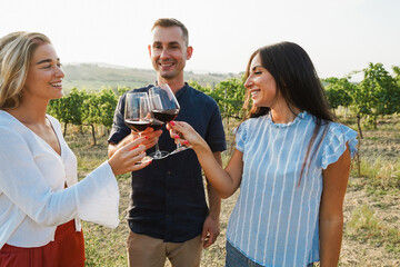 Naklejka premium Happy adult friends having fun drinking red wine with vineyard in background - Three people doing wine taste at summer time in countryside resort - Focus on faces