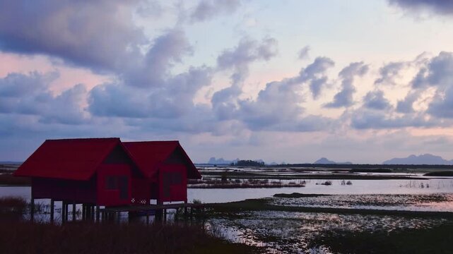 Timelapse zoom in Thale Noi Twin Wooden Shelters standing over reflective Phatthalung wetlands at sunrise, featuring iconic red-roofed stilt architecture and long Ekachai Bridge in rural Thailand.