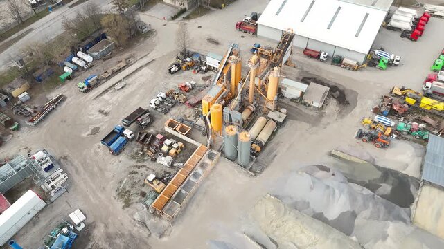 Aerial view of industrial enterprise showing sand and gravel piles, machinery, and trucks in a construction site, with buildings and cityscape visible in the background