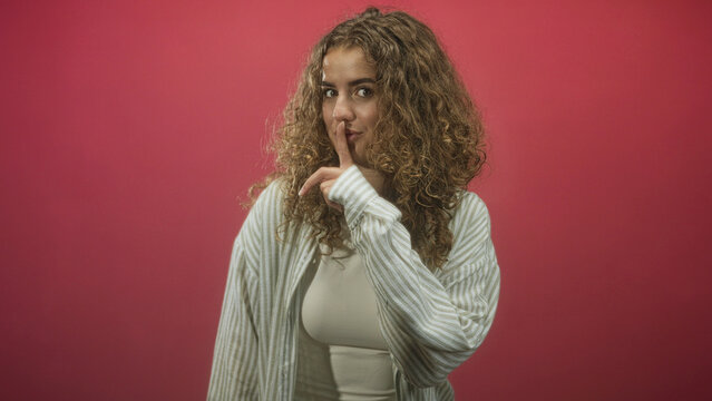 Young curly haired woman finger to lips gesture showing finger and upper chest, wearing striped shirt and tank top in studio; playful secrecy.