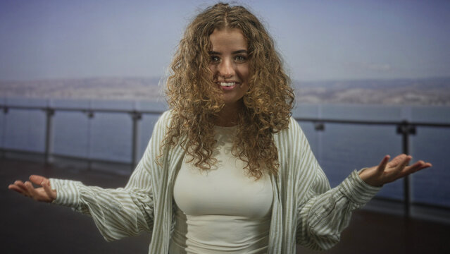 Woman with curly hair smiling, arms outstretched with palms up on building promenade railing by the sea; joyful openness.