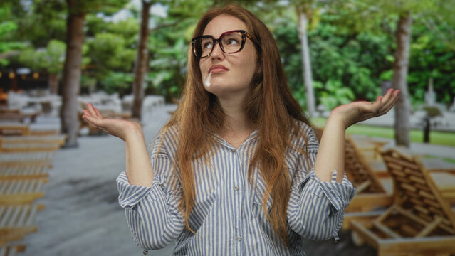 Woman raises hands under bright green foliage in forest clearing; confusion doubt uncertainty frustration.