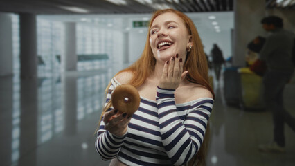 Fototapeta premium Redhead woman holding a glazed donut, hand covering mouth in a busy airport terminal with travelers and bins visible; playful delight.