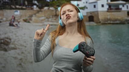 Young woman wearing cat ear headphones holds gamepad and points finger in front of seaside building while pouting and squinting in displeasure  frustration defiance. © Krakenimages.com