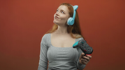 Woman holding a gamepad and wearing blue cat ear headphones, hand gripping a turquoise accented controller against a red studio wall  playful joy. © Krakenimages.com
