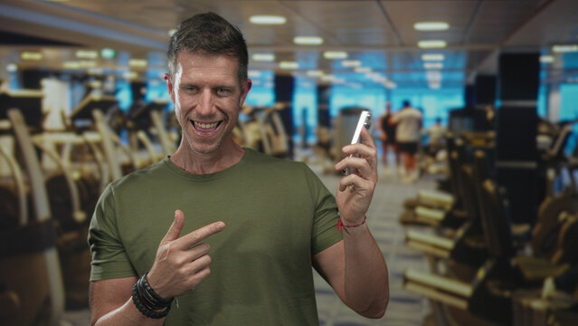 Man points finger to smartphone in building gym while smiling and showing hand; confidence connection.