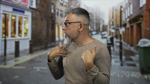 Man points thumbs to chest on a city street wearing glasses and brown sweater; surprise self reference.