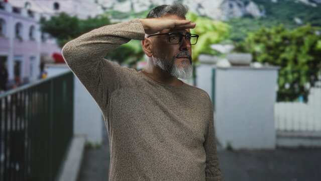 Man smiling with hand shielding eyes on street wearing glasses and sweater in casual pose by railing and building; contentment reflection.