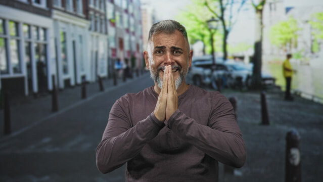 Man with hands pressed together in a pleading gesture on a city street showing beard and casual shirt; desperation plea.
