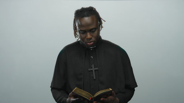 Young african american priest in black cassock reading a gilded book, holding it with hands in a pale gray studio; faith contemplation devotion.
