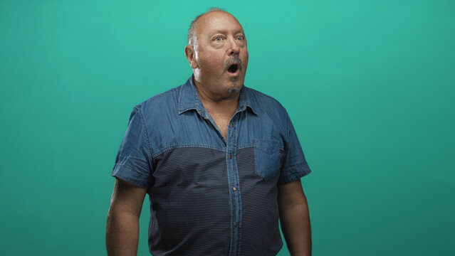 Senior hispanic man with open mouth gasp and visible bare neck wearing denim shirt, turned profile against teal studio backdrop; surprise reaction.