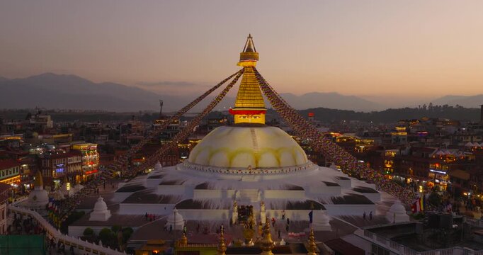 Pull in aerial view Boudhanath Stupa, UNESCO World Heritage Site in Kathmandu Nepal, moving from the glowing monuments to the illuminated pinnacle with prayer flags and city at dusk.