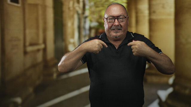 Man points fingers to chest while wearing glasses and black polo in a stone building corridor; surprise self recognition.
