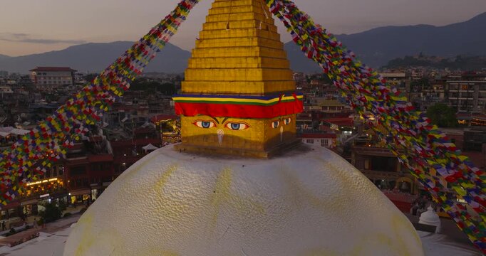Aerial revealing from the eyes of peace of Boudhanath Stupa, a UNESCO World Heritage Site, Kathmandu Nepal, expanding to show the full, prayer flags and surrounding city during sunset golden hours.