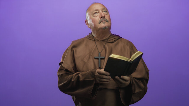 Man monk holding a bible with hands while reading, wearing brown robe and wooden cross, posed against purple backdrop in studio; serenity contemplation.