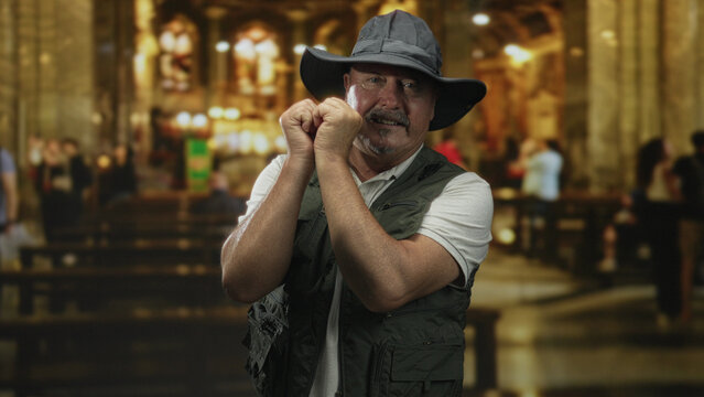 Senior hispanic fisherman in vest and hat, hands clasped near face in ornate cathedral building; reverence quiet reflection.
