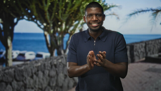 Man gesturing with hands and smiling while wearing glasses and a navy polo on a seaside street with stone wall and palm trees; joy presentation.