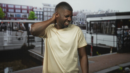 Fototapeta premium Man holding neck for backache and grimacing on street by a canal boat and dock, with nearby buildings and pavement visible; discomfort.
