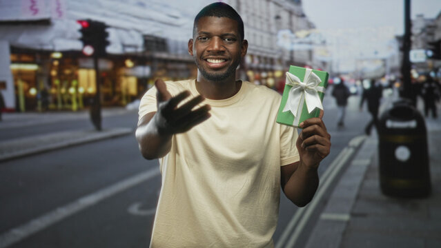 Man holding green gift box with white bow, smiling and reaching out left hand while standing on a busy city street offering a present; generosity welcome.