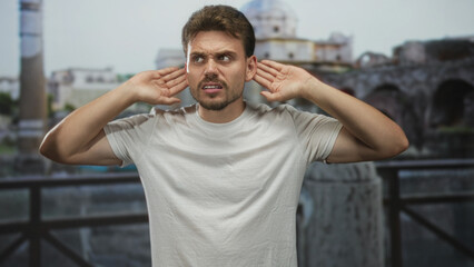 Fototapeta premium Man with hands cupping ears at building ruins on an outdoor balcony, looking upward and tensing shoulders; frustration focus.