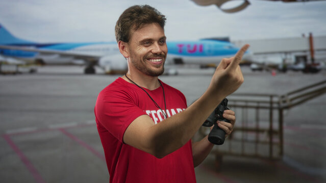 Lifeguard man smiling pointing finger at airport tarmac wearing red shirt and holding binoculars; vigilance duty preparedness.