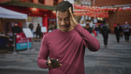 Naklejka premium Man holding smartphone with hand to head on street market, wearing maroon shirt and checking phone with a pained expression; frustration distraction.