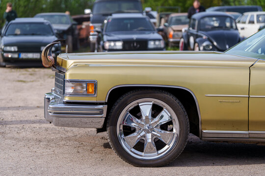 Side profile of a gold Cadillac Fleetwood with custom bull horns on the hood and chrome rims, classic American luxury car at an outdoor automotive show