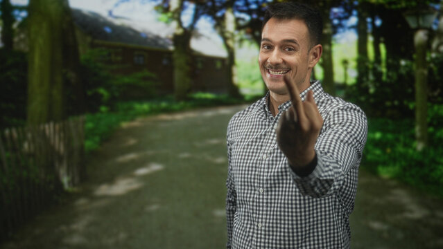Man with moustache and checkered shirt showing middle finger and smiling while standing on a forest path near a cottage; defiance.