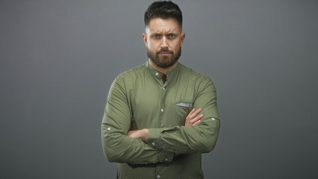 Bearded man with arms crossed and furrowed brow wearing green button shirt in studio; reserved skepticism caution.